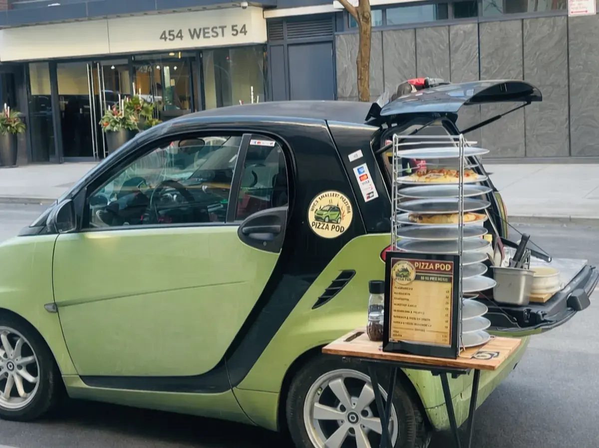 The green Pizza Pod Smart Car at a Midtown curb with its tailgate open, a tall rack of pie pans at the back and the menu board propped on a table