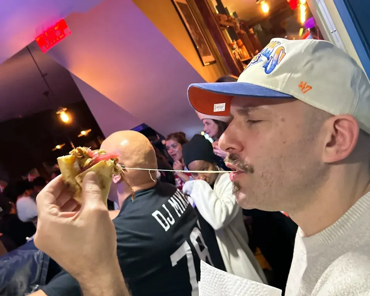 A guest pulling a slice at a late-night party, the cheese stretching, a crowd behind him under coloured light