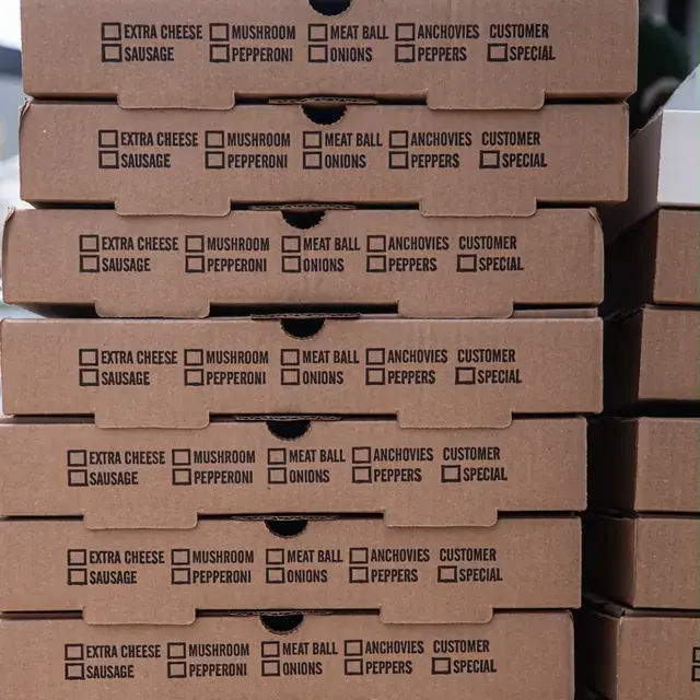 A wall of stacked kraft pizza boxes, each printed with a row of topping tick-boxes