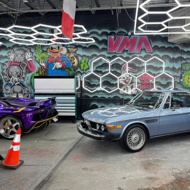 A vintage BMW and a purple supercar in a mural-covered garage under hexagon lights