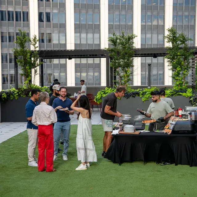 Guests with plates at the black-draped service table on the rooftop lawn, a cook serving behind it and office towers over the pergola