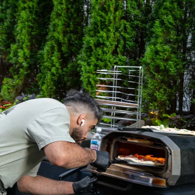 A cook bending to check a pie in a countertop oven, a tall pie rack and a clipped hedge behind