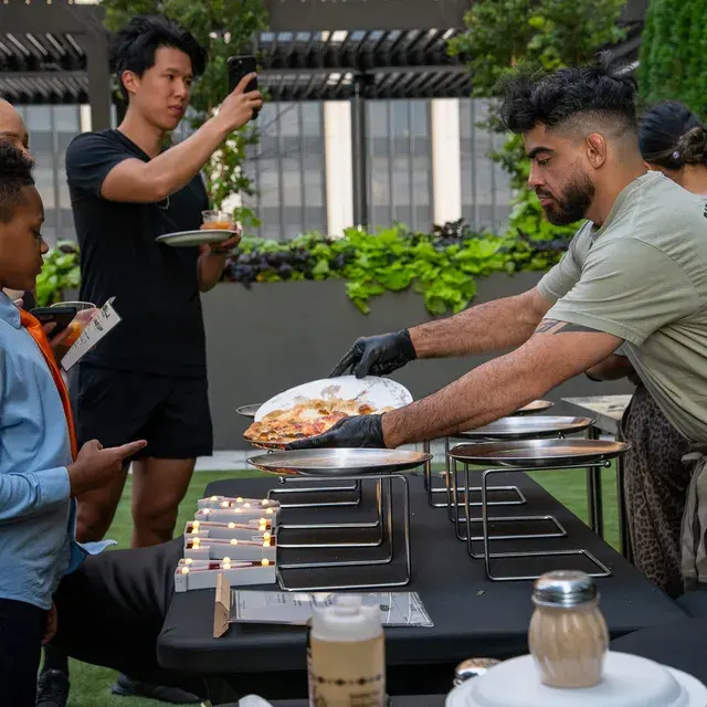 A cook lifting a finished pie onto a raised stand while a guest takes a photo of the table
