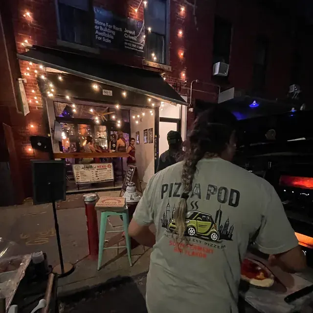 A cook in a Pizza Pod shirt with a pie on a peel outside a lit storefront at night