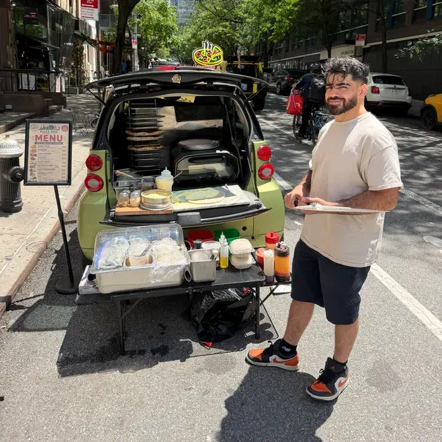 The Smart Car at the curb with its tailgate open on a pie rack and an oven, a cook and the menu board beside it