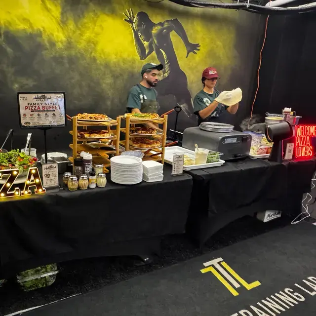 A family-style pizza buffet laid out on black-draped tables against a yellow mural, two cooks working the line behind it