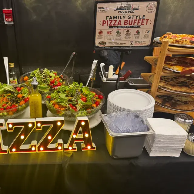 Light-up PIZZA letters beside bowls of salad and a rack of finished pies on the buffet table