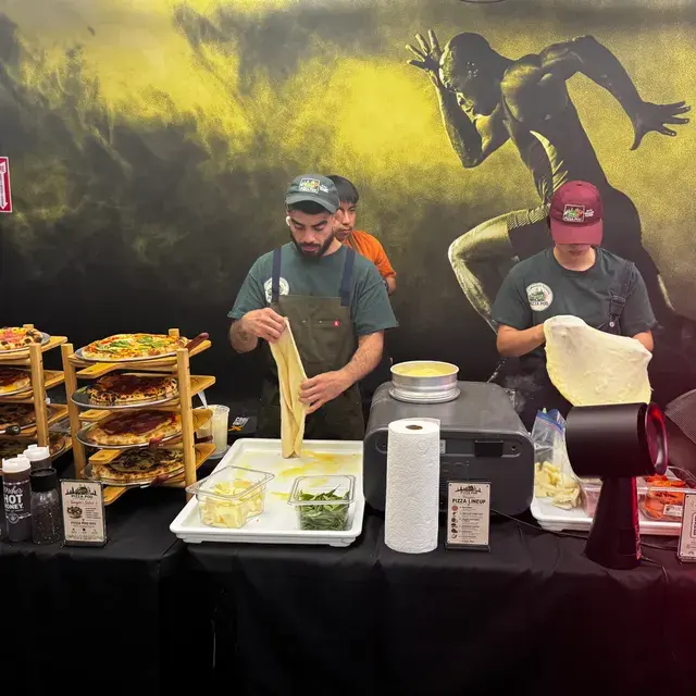 Two cooks stretching dough side by side behind the buffet, trays of prepped toppings in front of them