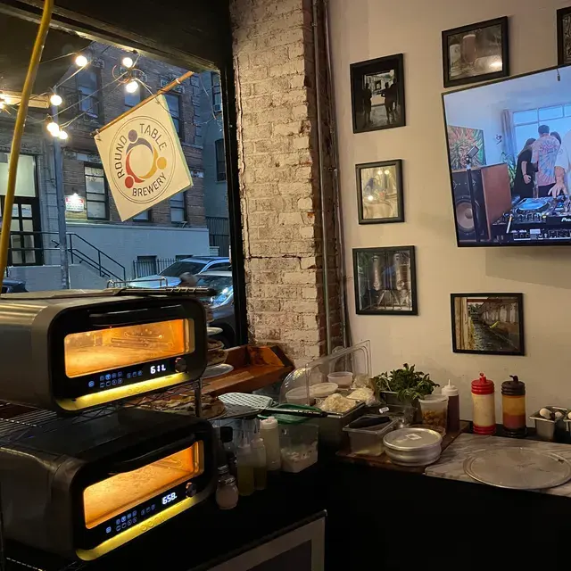 Two ovens glowing at the front window under the Round Table Brewery flag, beside framed pictures and a screen
