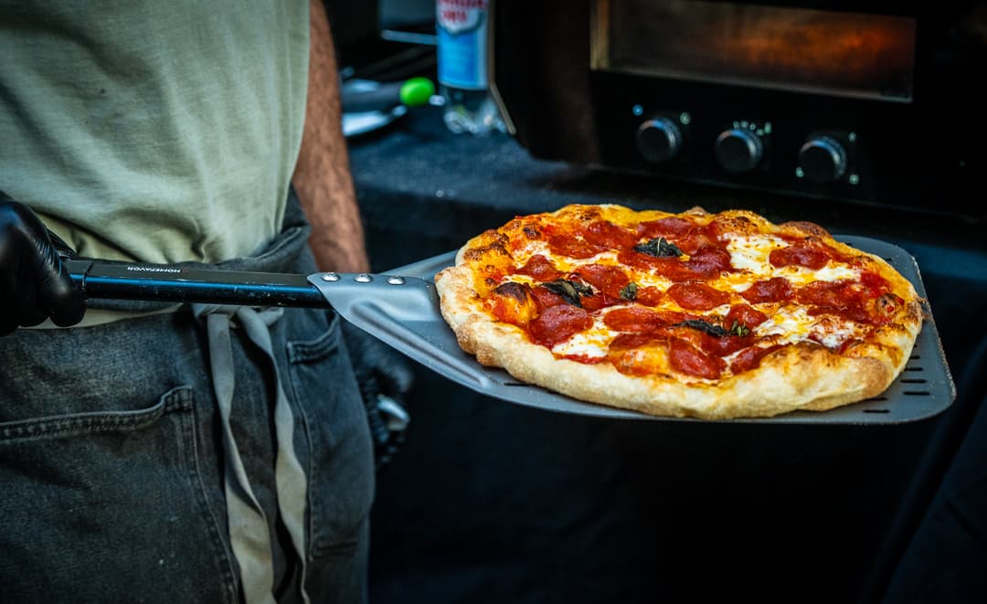 A pepperoni pizza on a peel, held in front of the oven's dials.